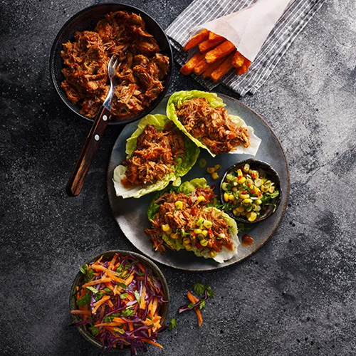 Slimming World food range barbecue pulled pork in lettuce cups on a grey plate. Sweet potato fries on the side. All placed on a grey table.