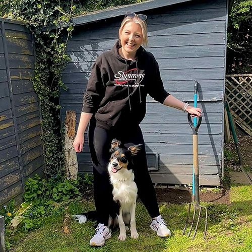 Slimming World Consultant Katie holding a digging fork in her garden. Her dog is sitting between her legs.